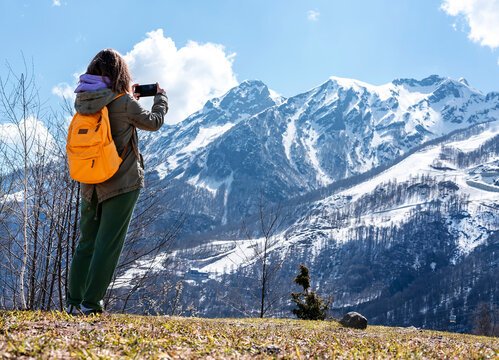 Young woman tourist with yellow backpack taking photo on smartphone of snow covered mountain range against blue sky using modern technology beauty in nature, walking photographing