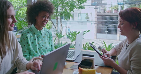 Three diverse young women using modern technology and talking at coffee shop table. Group of female friends in conversation while working remotely at cafe restaurant