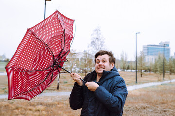 Shocked European white young male man fool around with umbrella in city park, strong storm wind,...