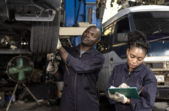 Black Mechanic Woman Listing Car Problems On Clipboard With Black African Mechanic Man Fixing Car Background, Car Mechanic Concept