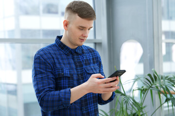 Portrait of happy handsome serious guy, young European blonde man in shirt is using his cell mobile phone, looking at screen of smartphone, reading or typing message in office building indoors.