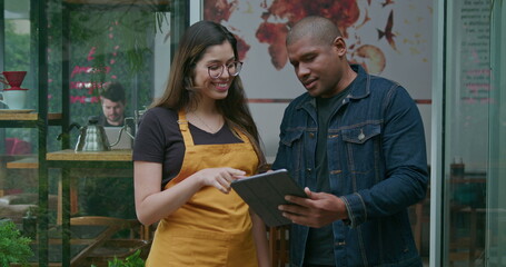 An African American manager of small business holding tablet speaking with female employee at coffee shop entrance. Portrait of two diverse happy staff of restaurant cafe place