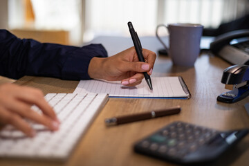 Woman types on the keyboard and writes notes on a page. Desk with calculator and office implements. Calculating, accounting, clerk, financial business and back office.