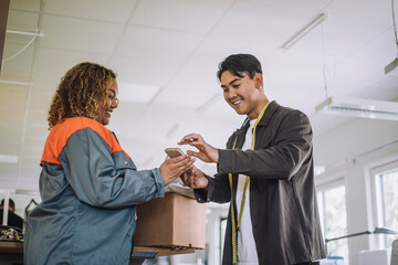 Smiling male fashion designer giving digital signature while receiving package from delivery person at workshop
