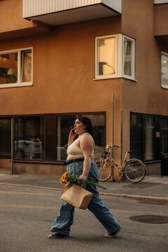 Side view of voluptuous woman talking on mobile phone while walking with shopping bag on road