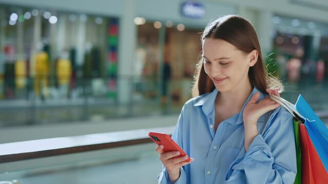 Smiled female customer holding her purchase in the paper bags going on the mall. Lady carrying her shopping-bags, using her smartphone. Woman after shopping, texting