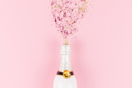 A Christmas tree bauble in the shape of a white champagne bottle. Golden and pink glitter come out of the bottle like a firework.
