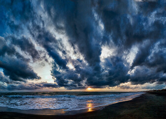Fantastic stormy sky with stratocumulus clouds at dramatic weather panoramic view over the beach , impressive blue hour with golden sunbeams hits rough foamy threatening sea waves crashing at shore