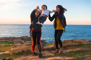 Aunt of the child and mother playing with him child on the coast by the sea at sunset