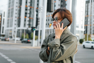 Fototapeta premium Stylish young asian woman with short haircut and glasses talking on phone on city street. Side view portrait of young woman calling by smartphone outdoors