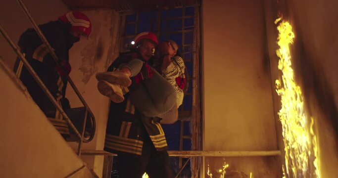 Brave Fireman Descends Stairs of a Burning Building and Holds Saved Girl in His Arms. Open fire and one Firefighter in the Background.