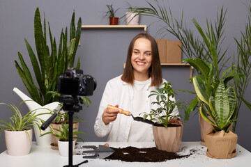 Indoor shot of smiling positive woman blogger florist sitting at table in her greenhouse in front...