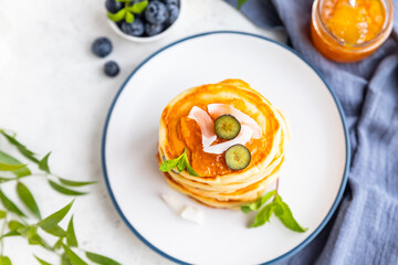 Stack of fluffy pancakes with orange jam, blueberries, coconut chips and mint, light background. Traditional breakfast. High key photography.