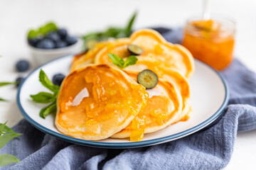 Traditional fluffy pancakes with orange jam, blueberries, coconut chips and mint for healthy breakfast, light background. High key photography.