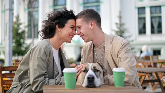 A happy couple outdoors near a cafe. Petting their dog and looking at each other, coffee on the table. Autumn atmosphere. Slow motion