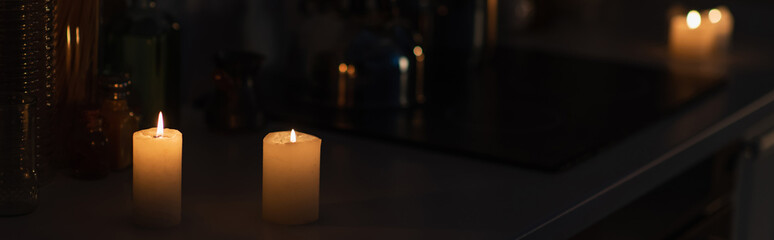 candles burning on kitchen worktop in darkness during energy blackout, banner.