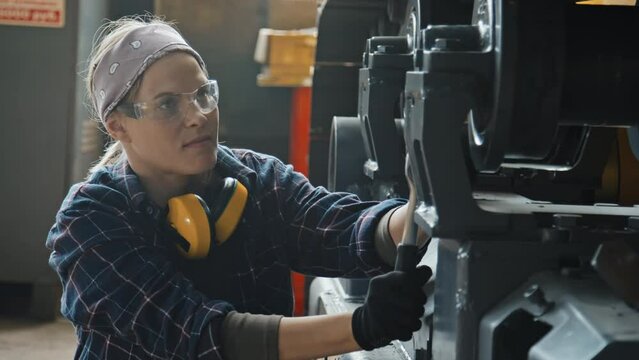 Female service technician in safety glasses using bolt wrench while fixing wheel of tractor machine at plant