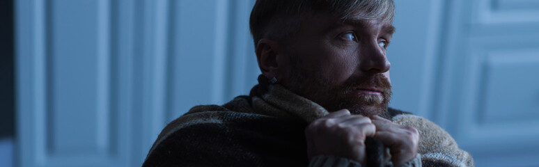 upset man covered with warm blanket looking away at home during energy blackout, banner.