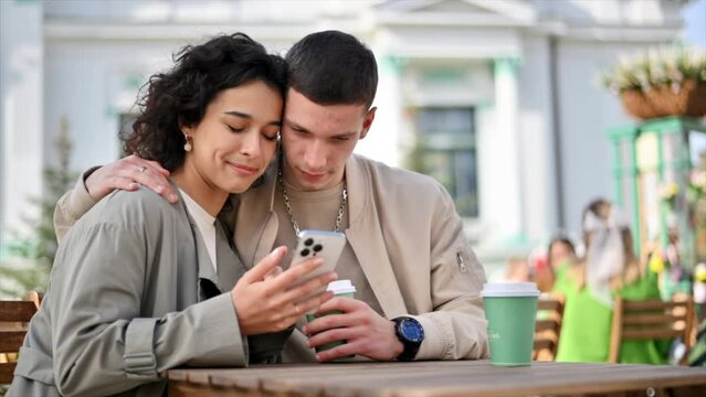 A happy couple outdoors near a cafe. Man is hugging his woman, looking in the phone, smiling, talking, coffee. Autumn atmosphere