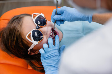 A girl of European appearance in funny smile goggles receives dental treatment with smile lying in an orange chair in a dental clinic