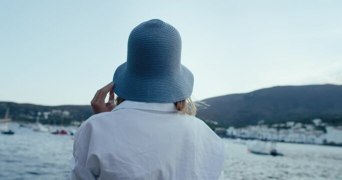 Portrait of authentic and real young woman tourist in bohemian white outfit and blue bucket hat look at camera, smile, turn around to enjoy view on holiday village on mediterranean coast