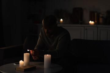 man lighting candles in dark kitchen near mobile phone and book during energy blackout.