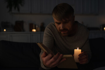 man holding lit candle while reading book in dark kitchen during electricity outage.