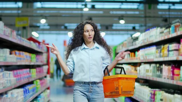 cheerful young woman customer in the store dances funny between the rows in a supermarket with a basket in his hands. walking the grocery store and moving like dancing while doing shopping having fun