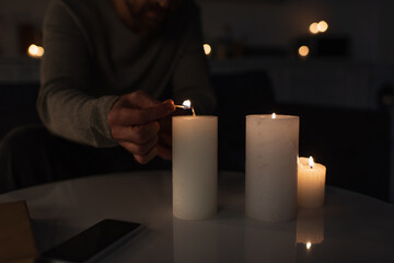 cropped view of man in darkness lighting candle with burning match near smartphone with blank...