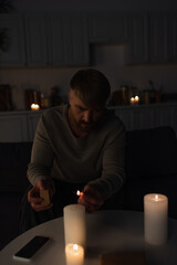 man sitting in dark kitchen during energy blackout and lighting candles.