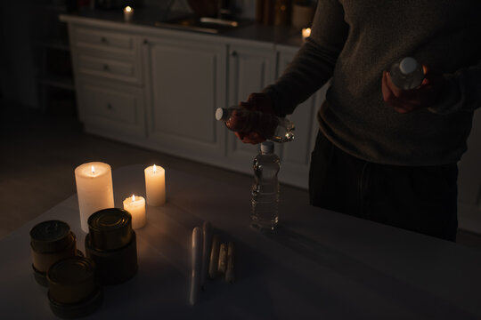 cropped view of man with bottled water near table with canned food and candles in dark kitchen.