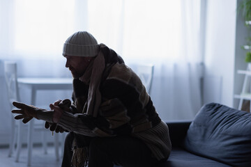 side view of frozen man putting on gloves while sitting on couch in hat and warm blanket.