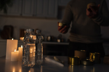 reserve of bottled water and canned food with candles near cropped man in dark kitchen.