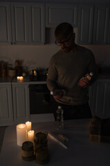 man in eyeglasses holding bottled water near reserve of canned food and candles on table in dark...