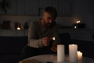 man holding burning match while lighting candles in dark kitchen during power outage.