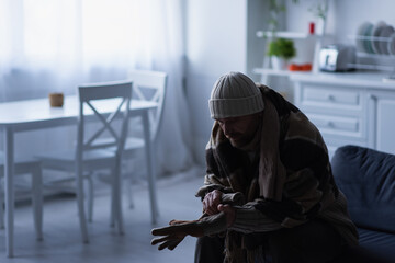 frozen man in knitted hat sitting in kitchen under blanket and wearing warm gloves.