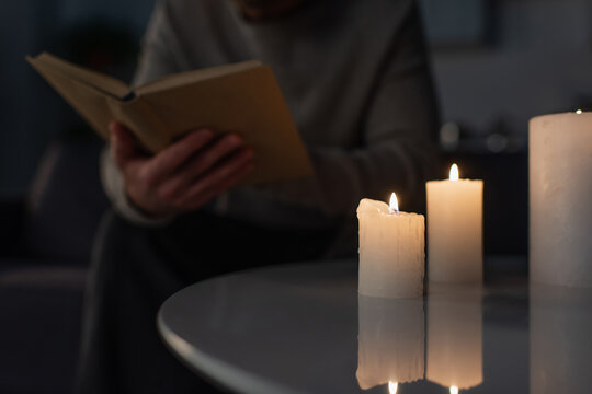 partial view of blurred man reading book in darkness near burning candles on table.