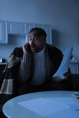 shocked man under warm blanket holding invoices and talking on smartphone during power outage.