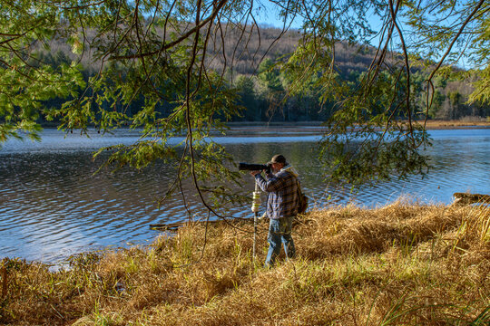 A male photographer using his monopod and long lens stands on the edge of a lake taking photos of the far shoreline at Hawkins Pond Park in Broome County in Upstate NY.