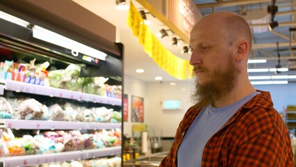 Bearded man looks at shelves with food in supermarket, look over assortment and chooses products. Man buys fresh and tasty food for dinner at grocery store. Thinking about proper and healthy nutrition