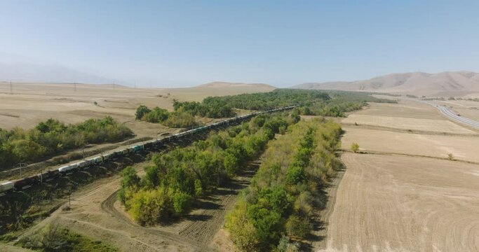 Freight long cargo train carries lorry carriages by arid hilly landscape. Railways in pampas. Aerial drone perspective view at summer sunset