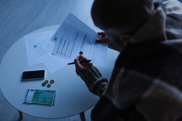 high angle view of man holding invoices near money and mobile phone in twilight.