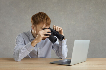 Young business man freelancer office employee looking at laptop screen through binoculars searching content on internet sitting at desk. Project idea find or job search online concept. Studio shot