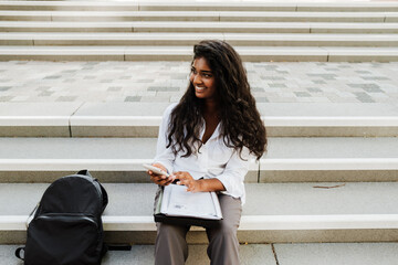 Young indian woman using cellphone while sitting at stairs outdoors