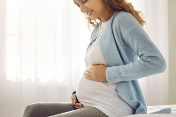Crop of smiling healthy Caucasian pregnant woman sit on sofa relax on maternity leave at home. Happy young female excited with pregnancy wait for childbirth. Motherhood and parenting.