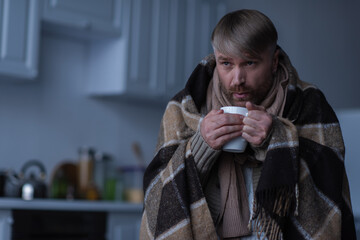 man standing under warm blanket and blowing at hot tea during power outage at home.