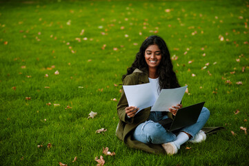 Brunette woman working with laptop and papers while sitting on grass
