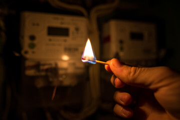 A man's hand in complete darkness holds a burning match to read the home electricity meter.