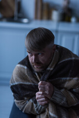 displeased and frozen man looking away while sitting under warm blanket during power blackout.