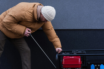 side view of man in warm jacket and knitted hat starting power generator outdoors.